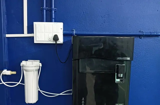 Black wall-mounted RO water purifier with a pre-filter housing on a blue wall